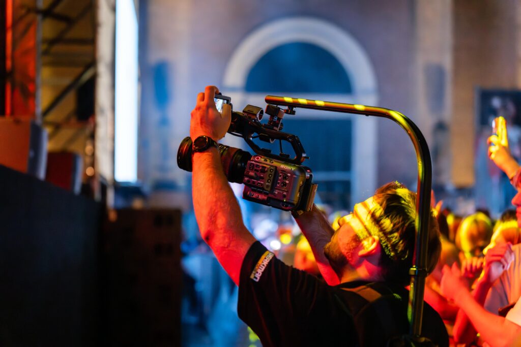 Videograaf met Sony cine-camera legt kleurrijk indoor live-evenement vast voor dynamische social media content, omringd door stage-verlichting en enthousiast publiek.