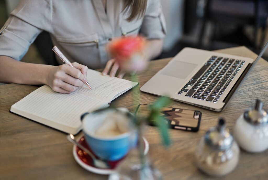 Mastering the First Impression: Your intriguing post title goes here Woman writing in a notebook with a laptop and coffee cup on a desk. Ideal for workspace inspiration.